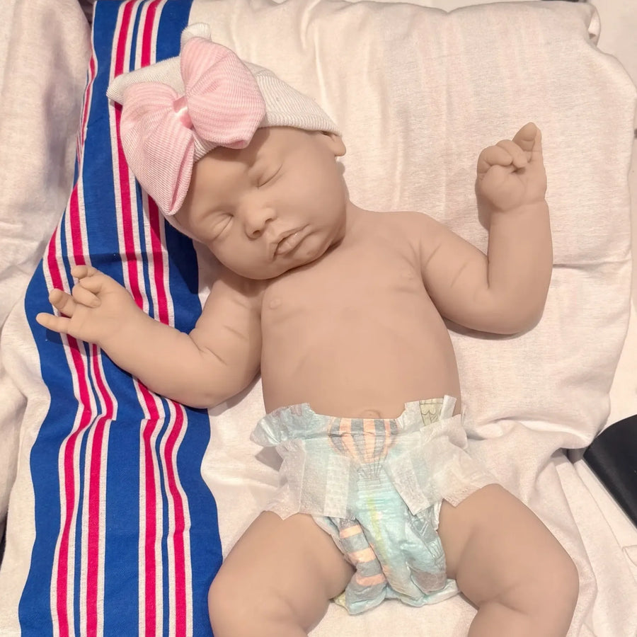Newborn baby doll wearing a pink headband and colorful diaper on a striped blanket.